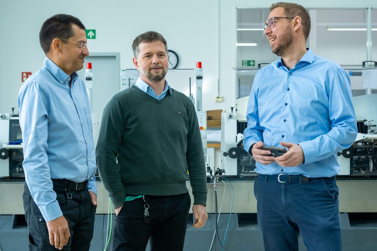 Khalid Dawam, Jan Schölzel und Ronny Richter stehen in einer modernen Werkstatthalle von PostPrint Leipzig und unterhalten sich. Sie tragen Hemden oder Pullover und wirken entspannt und freundlich. Im Hintergrund sind Maschinen und Arbeitsmaterialien zu sehen.
